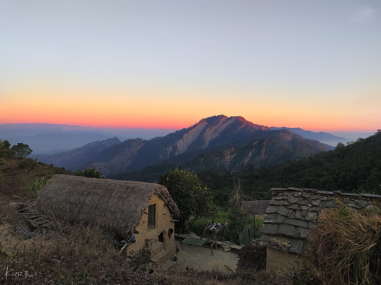 Thatched village houses below forested hills under a pastel sunset sky